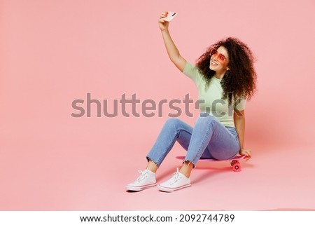 Full size body length young curly latin woman 20s wears casual clothes sunglasses sit on skateboard do selfie shot on mobile cell phone isolated on plain pastel light pink background studio portrait
