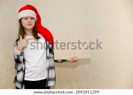 A girl in a Santa Claus hat with a doubt on her face points to a cardboard plate with a place for the text. Idea - I can't believe my eyes that there are such discounts