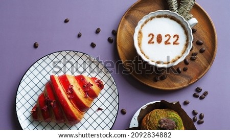 Holidays food and drink theme coffee cup with number 2022 over frothy surface served on wooden plate with dried pine branches flat lay on purple background with coffee beans, croissant, cruffin.
