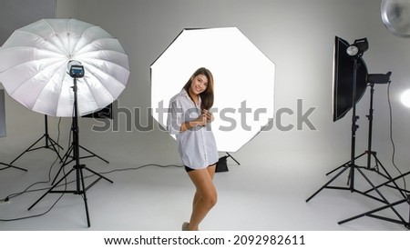 Asian young happy cheerful female model in plaid dress standing smiling look at camera posing in photographing studio shooting set full of softbox lighting reflection on white backdrop background.