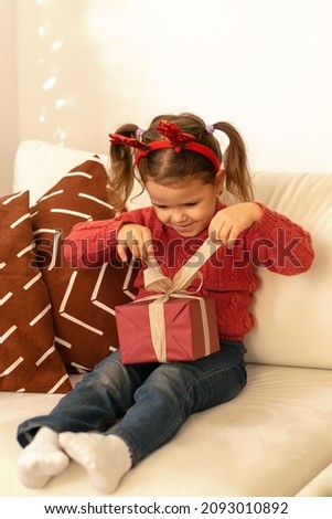 Little caucasian girl unpacks gift boxes at home.The girl is wearing a red sweater and jeans.Girl has a festive headband with deer horns on her head.New Year,Christmas concept.Selective focus.