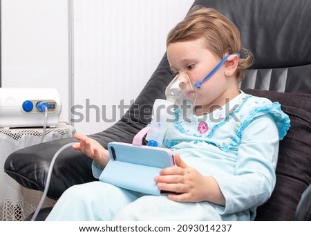 A Caucasian child, a girl of 5 years old, watches cartoons on a smartphone and does preventive maintenance of the respiratory tract, therapeutic inhalation.