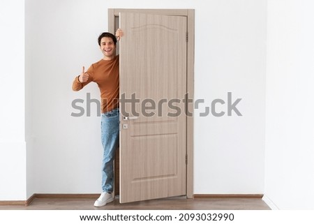 Good Offer. Portrait of cheerful young guy standing in doorway of his apartment, smiling millennial male homeowner holding ajar door looking out, greeting visitor, showing thumbs up like sign gesture