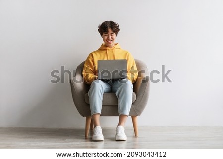 Happy young Asian male working online, sitting in armchair and using laptop against white studio wall, full length. Joyful millennial man surfing internet, watching webinar on portable computer Royalty-Free Stock Photo #2093043412