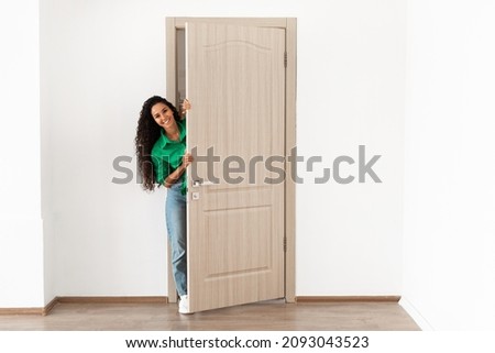 Portrait of cheerful young woman standing in doorway of modern apartment, millennial female homeowner holding slightly open ajar door looking out and smiling, greeting visitor, full body length Royalty-Free Stock Photo #2093043523