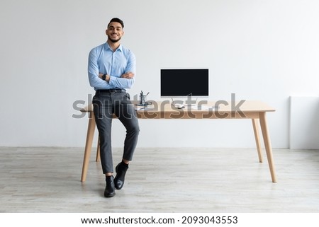 Mock Up Template. Happy Smiling Arab Man Sitting On Desk Posing With Folded Arms Showing Pc Computer With Blank Black Screen At Home Office, Free Copy Space. People, Technology, Remote Work Concept Royalty-Free Stock Photo #2093043553