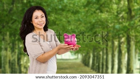 Composite image of asian senior woman holding a piggy bank against tress in park. savings concept