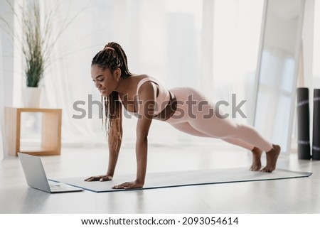 Full length of sporty young black woman standing in plank, training to online sports video on laptop at home. Athletic African American lady exercising abs muscles, doing domestic workout Royalty-Free Stock Photo #2093054614