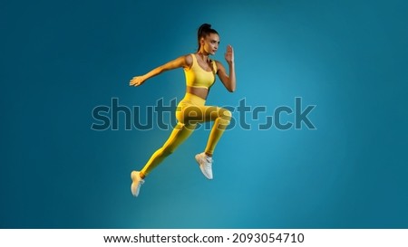 Motivated Young Lady Jumping Posing In Mid-Air Over Blue Studio Background, Looking Aside. Fitness Woman Exercising Wearing Yellow Sportswear Running In Air. Panorama, Full Length Shot Royalty-Free Stock Photo #2093054710