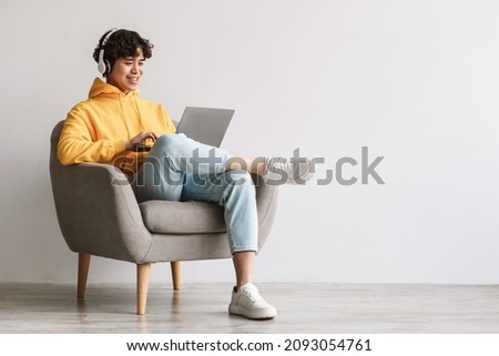 Happy young Asian man using laptop, wearing headphones while sitting in armchair against white wall, copy space. Millennial male communicating online, working or studying remotely on portable pc Royalty-Free Stock Photo #2093054761