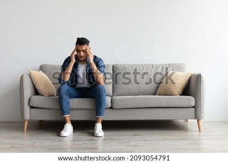 Sad young Middle Eastern man suffering from headache or migraine, touching head massaging temples. Stressed guy sitting on couch with painful face expression feeling terrible weakness or depression Royalty-Free Stock Photo #2093054791