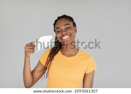 End of coronavirus pandemic. Happy black woman taking off medical mask, feeling free on grey studio background. African American female removing facial covid protection, celebrating end of lockdown Royalty-Free Stock Photo #2093061307