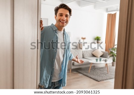 Portrait of cheerful man welcoming inviting visitor to enter his home, happy young guy standing in doorway of modern apartment, millennial male holding door looking out showing living room with hand Royalty-Free Stock Photo #2093061325