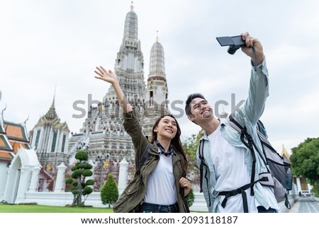 Portrait of Asian couple travel in city for honeymoon trip at morning. Attractive young man and woman backpacker tourist, enjoy spend time on holiday vacation together and using mobile phone selfie.