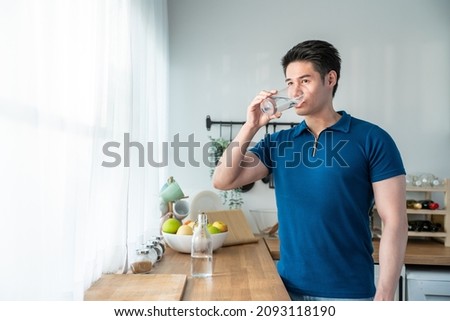 Asian attractive young man stand drinking water and looking out window. Handsome male drink or take of mineral natural in glass after wake up in morning for health and wellbeing in kitchen in house. Royalty-Free Stock Photo #2093118190