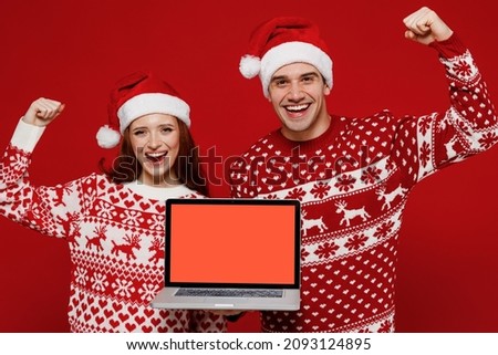 Young couple friends two man woman in sweater hat hold use work on laptop pc computer blank screen workspace area do winner gesture isolated on plain red background studio. Happy New Year 2022 concept