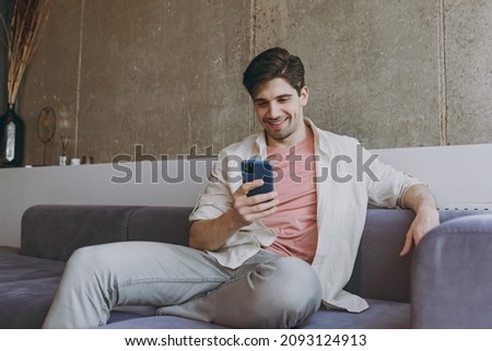 Young smiling happy man 20s wearing casual clothes beige shirt pink t-shirt use mobile cell phone chat online browsing sitting on grey sofa rest indoors at home on weekends. People leisure concept. Royalty-Free Stock Photo #2093124913