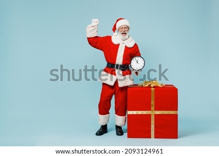 Full body old Santa Claus man in Christmas hat red suit clothes stand near big gift box hold clock do winner gesture isolated on plain blue background studio Happy New Year 2022 merry ho x-mas concept