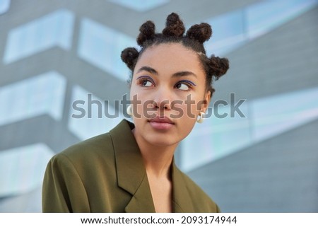Stylish young woman with hair buns concentrated away with serious expression wears vivid blue eyeliner dressed in green stylish jacket poses against blurred background. Street lifestyle concept Royalty-Free Stock Photo #2093174944