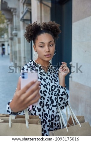 Vertical shot of thoughtful curly haired young woman takes selfie on modernsmartphone carries shopping bags with purchases dressed in fashionable blouse poses against blurred background outdoors.