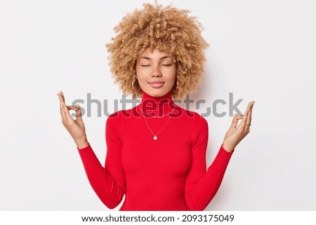 Calm concentrated woman with curly hair keeps eyes closed and meditates indoor makes mudra gesture wears casual red turtleneck isolated over white background tries to relief stress. Zen pose