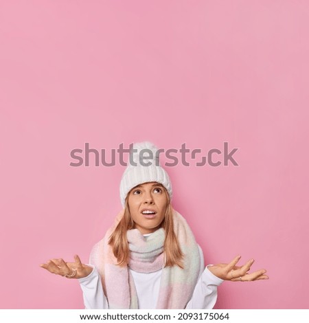 Indignant indifferent woman spreads palms feels hesitant and clueless focused overhead wears winter hat and scarf around neck poses against pink background copy space for your promotional content
