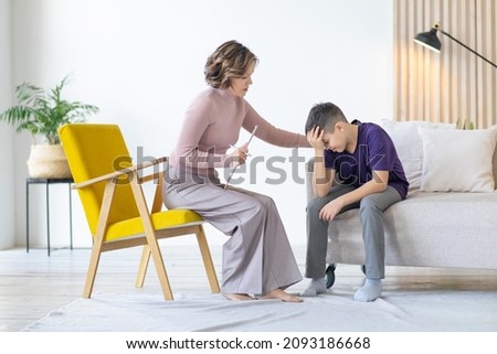 Counseling adolescent psychologist calms and holds adolescent by shoulder. Boy in purple t-shirt is worried and holds his head with his hand, while sitting on couch in minimalistic room. Royalty-Free Stock Photo #2093186668