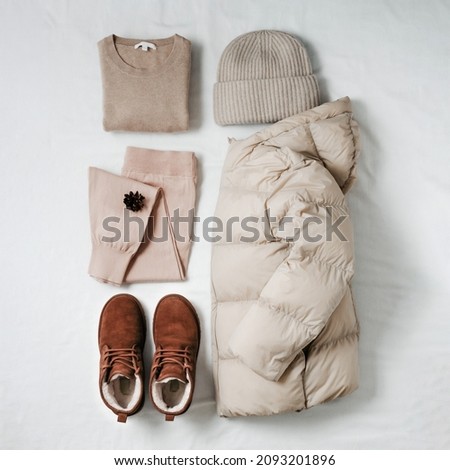 Women warm puffer light down jacket, beige cashmere sweater, pink knitted joggers, knitted bini hat, brown fur chukka suede boots on white bed sheet. Cozy winter clothing knolling in beige monochrome Royalty-Free Stock Photo #2093201896