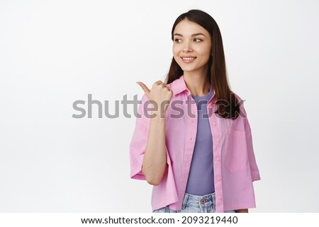 Young female student, pointing and looking left with hopeful smiling face expression, standing over white background