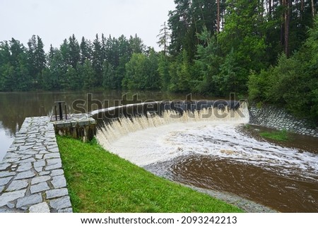 Landscape picture of the flooded old historical pond or water reservoir after a heavy rains with overflow waterfall and stream. Taken in southern bohemia region of Czech republic in summer rainy day.