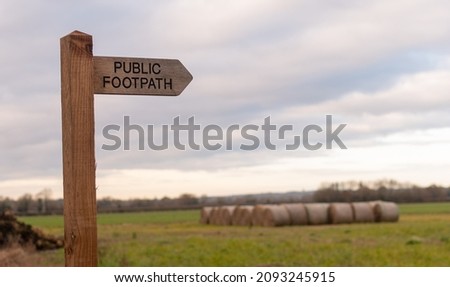 Wooden public footpath sign with blurred hay bales in background, UK countryside scene