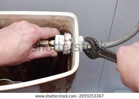 The plumber's hands unscrew the filling pipe nut from old dirty water fill valve in the empty drain water tank with wrench tool closeup - DIY WC repair at home Royalty-Free Stock Photo #2093253367