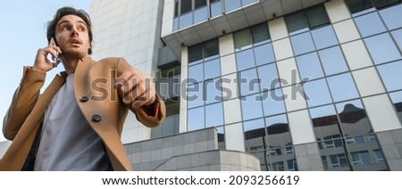 Low angle view of businessman in coat talking on mobile phone during wind near skyscraper outdoors, banner	 Royalty-Free Stock Photo #2093256619