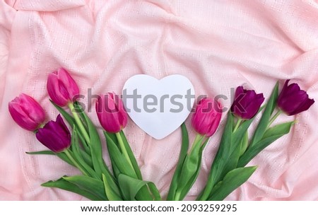 Pink, violet tulips on background of pink linen cloth with white paper card note in the shape heart with space for text. Top view, flat lay