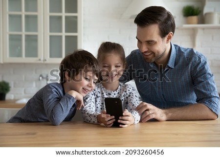 Happy Caucasian father with kids using modern smartphone together, sitting at table, smiling young dad with little son and daughter looking at phone screen, watching cartoons, making video call