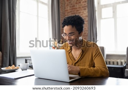 Happy young inspired African American woman in eyeglasses looking at computer screen, reading email with good news, enjoying studying or working distantly on online project at modern home office. Royalty-Free Stock Photo #2093260693