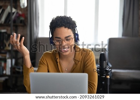 Happy pretty millennial African American woman in wireless headphones looking at laptop screen, enjoying video call meeting or voice acting using professional stand microphone, distant communication.