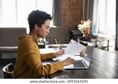 Focused serious young African American businesswoman manager employee in eyewear analyzing paper reports working on computer in modern office, preparing presentation or doing accounting paperwork. Royalty-Free Stock Photo #2093261035
