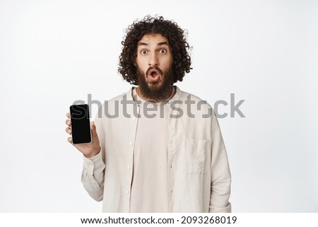 Image of impressed young middle eastern guy showing smartphone screen, saying wow, looking amazed, white background