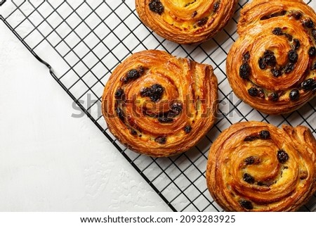 Just baked pain aux raisins on cooking wire rack. Buns are also called escargot or pain russe, is a spiral pastry with custard cream and raisin. Directly above, white table surface. Royalty-Free Stock Photo #2093283925