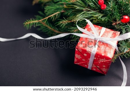Christmas and New Year gift box wrapped in festive paper and ribbon by fir wreath. Present for winter holidays on black background