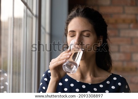 Peaceful young hispanic woman drinking glass of fresh pure water, preventing organism dehydration, keep dietary lifestyle, feeling thirsty standing near window at home, daily healthcare habit. Royalty-Free Stock Photo #2093308897