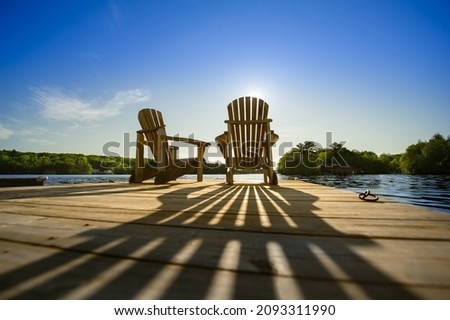 Cottage life - Sunrise on two empty Adirondack chairs sitting on a dock on a lake in Muskoka, Ontario Canada. The sun rays create long shadows on the wooden pier. Royalty-Free Stock Photo #2093311990