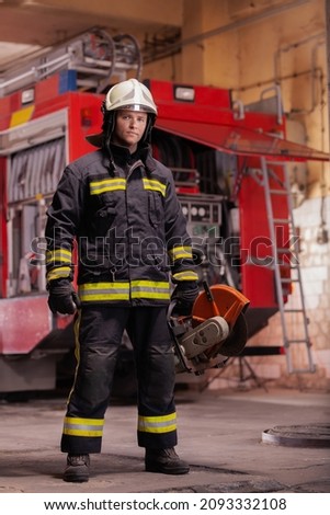 Professional firefighter in uniform wearing protective helmet and standing with emergency rescue equipment. Circular hydraulic cutting hard tool. Firetruck in the background.