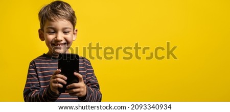 Front view portrait of small caucasian boy holding a smartphone in front of yellow background - little kid using mobile phone to take photos or play games while standing in studio with copy space