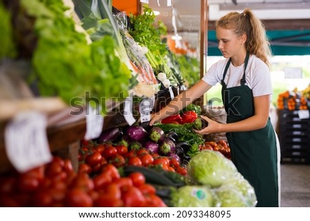 Young girl seller in uniform working in supermarket at her first job, checking organic eggplants Royalty-Free Stock Photo #2093348095
