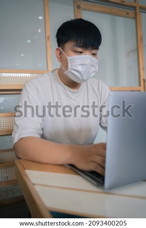 Focused young man wearing a medical mask using laptop, typing on keyboard, writing email or message, chatting, shopping, successful freelancer working online on computer, sitting in coffee shop.