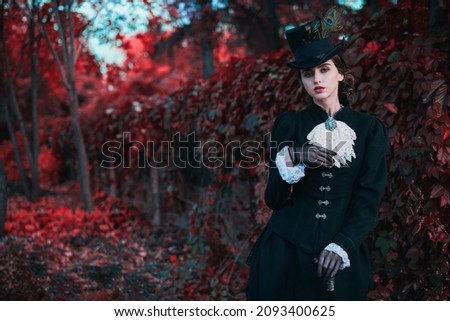 Historical reconstruction of the late 19th and early 20th centuries. Elegant brunette lady in a strict black dress posing in the background of red foliage. Historical makeup and hairstyle. Royalty-Free Stock Photo #2093400625