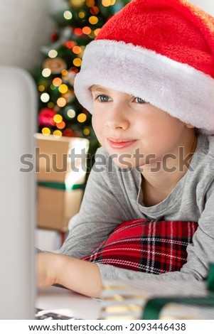 Preteen boy is typing on a laptop keyboard near the Christmas tree at home. Child is looking for gifts for the family in online store or watches a funny movie or cartoon during the winter holidays.