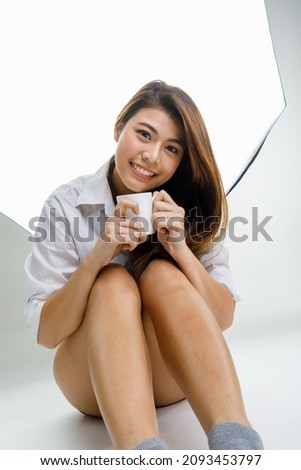 Portrait studio shot of Asian young happy cheerful female model in casual shirt standing smiling look at camera posing showing L fingers sign symbol in front big softbox lighting in photographing set.
